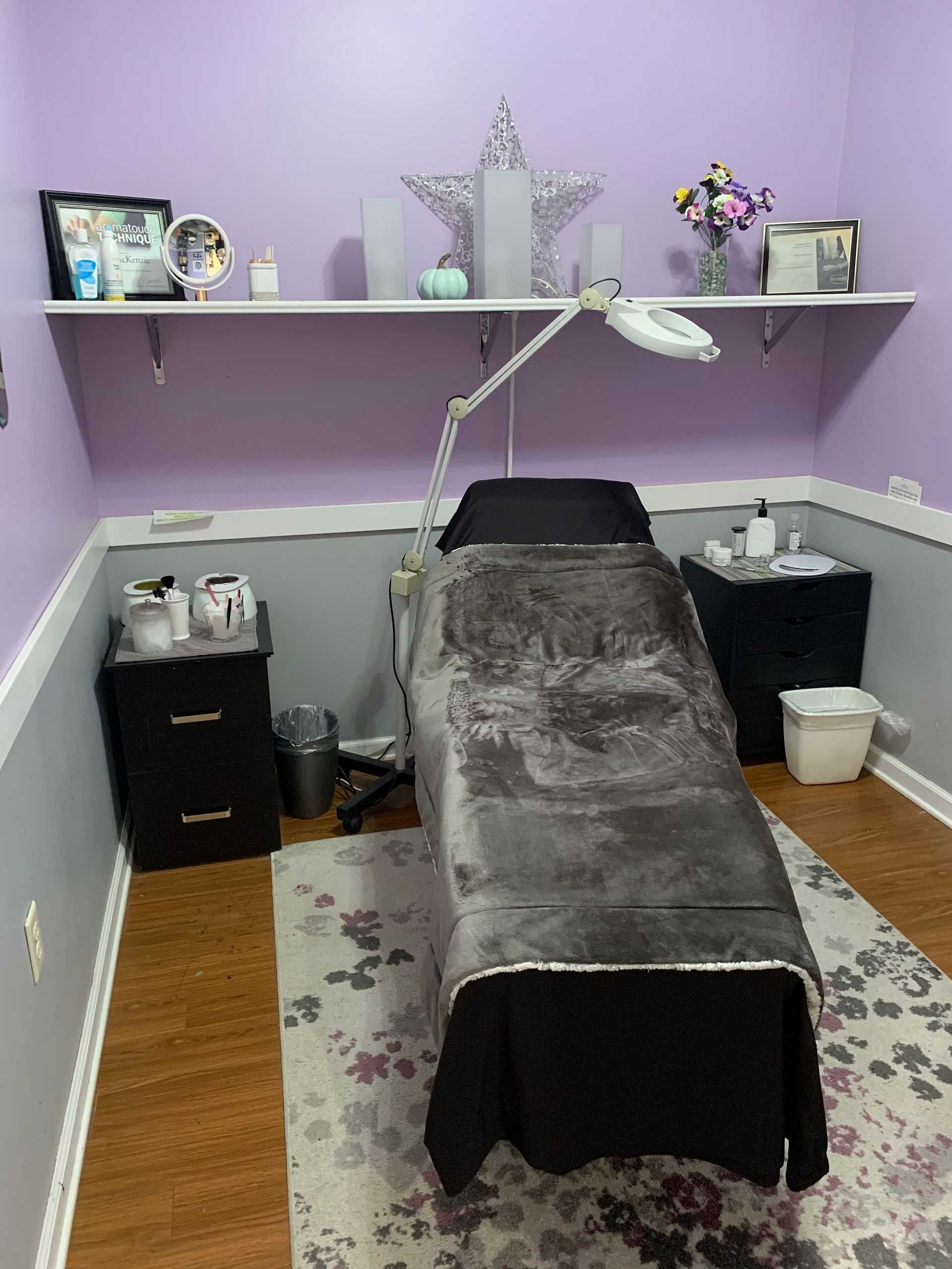 A cozy spa room with a treatment bed, purple walls, and a shelf with decorations and awards.